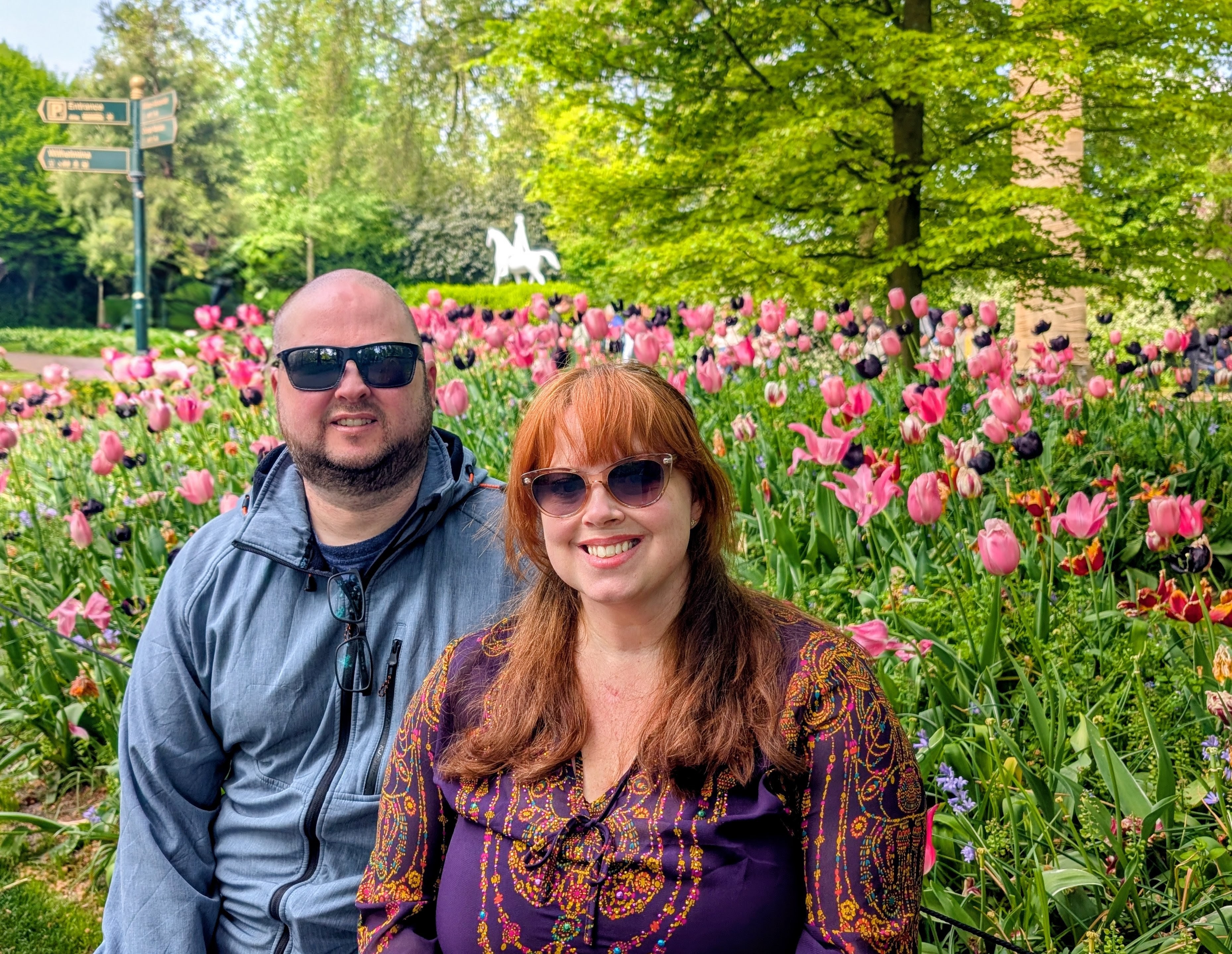 A man and a woman sitting in a garden filled with colorful tulips and greenery. Both are wearing sunglasses and smiling at the camera.